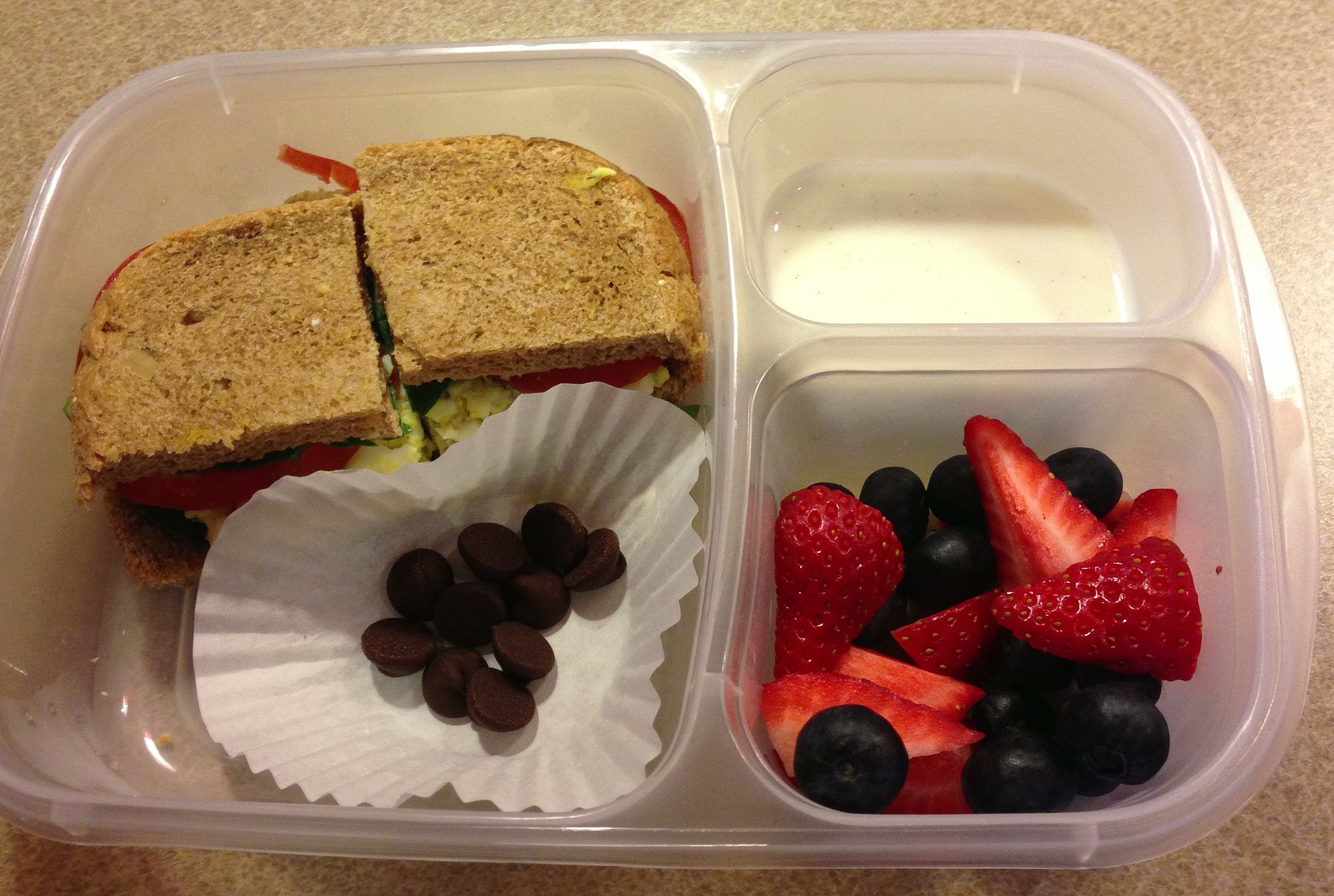 Egg salad on homemade multi-grain bread with spinach and tomatoes, strawberries and blueberries, vanilla bean yogurt and chocolate chips. All ingredients organic.